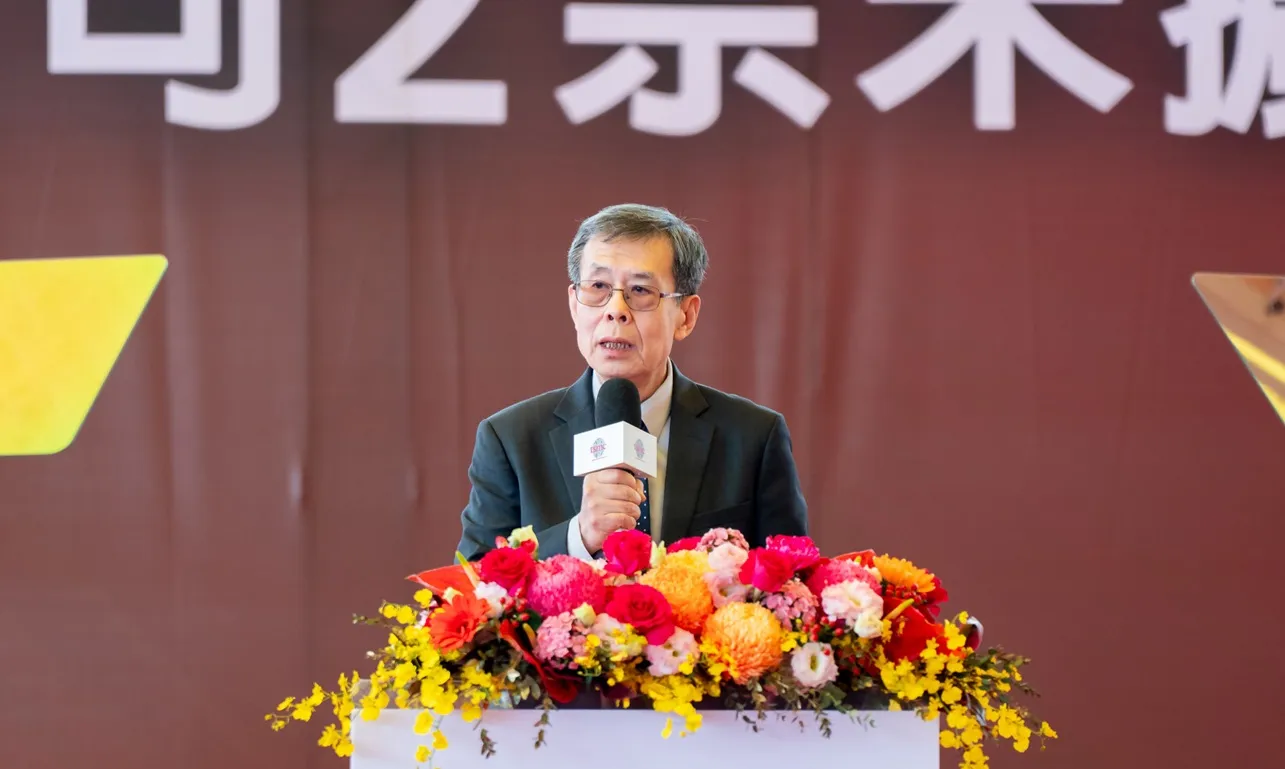 Y.P. Chyn, wearing a dark suit, speaks into a handheld microphone behind a floral arrangement.